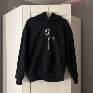 Svart hoodie med rosdetalj - Säljer en svart hoodie med en stilren vit ros på bröstet. Den är supermjuk och perfekt för kyliga dagar. Har en stor ficka fram och en skön huva. Passar både till vardags och mysiga hemmakvällar. Perfekt för dig som gillar enkel men snygg design!