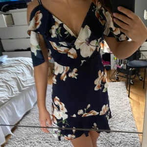 Blommig mörkblå klänning - Säljer en superfin mörkblå klänning med stora blommor i vitt och orange. Den har en omlott-design och offshoulder-ärmar som ger en romantisk look. Perfekt för sommarens fester eller en mysig vårdag. Klänningen är kort och har en volangdetalj vid fållen.