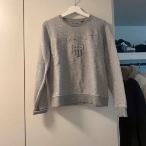 Grå sweatshirt från GANT - Säljer en stilren grå sweatshirt från GANT i storlek S. Tröjan har ett broderat GANT-logotyp på framsidan och är perfekt för både vardag och avslappnade tillfällen. Den är i bra skick och superbekväm att ha på sig. Passar perfekt till jeans eller joggers!