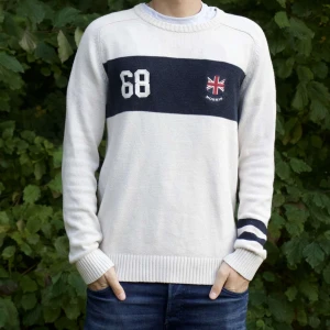 Beige tröja från Morris - Vi säljer en snygg beige tröja från Morris med en mörkblå rand över bröstet och siffran 68 samt en Union Jack-flagga. Tröjan har långa ärmar med mörkblå ränder på ena ärmen och ribbstickade muddar. Perfekt för en casual look!