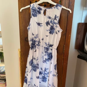 Vit och blå blommig klänning - Säljer en superfin vit klänning med blått blommönster och fjärilar. Den är ärmlös och har en luftig passform, perfekt för sommardagar. Klänningen har en liten knappdetalj i nacken och är gjord i ett lätt och bekvämt material. Perfekt för både vardag och festliga tillfällen!