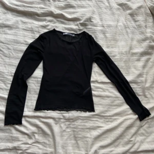 Svart långärmad topp - Säljer en stilren svart långärmad topp med vågiga kanter. Perfekt för en minimalistisk look. Toppen är i en mjuk och bekväm kvalitet.