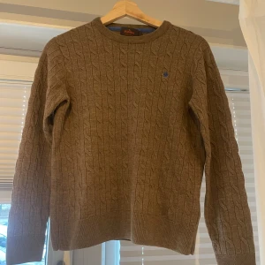 Brun kabelstickad tröja från Morris i 100% merinoull  - Säljer en stilren brun kabelstickad tröja från Morris i 100% merinoull. Tröjan har en diskret logga på bröstet och ribbade muddar vid ärmslut och nederkant. Perfekt för kyliga dagar!