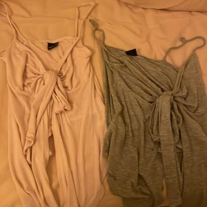 Två  blusar med knytning - Säljer två klänningar med knytning framtill. Den ena är ljusrosa och den andra är grå. Båda har smala axelband och en omlott-design som ger en elegant look. Perfekta för en somrig dag eller en avslappnad kväll.