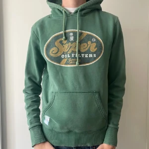 Super dry Hoodie - Säljer en grön hoodie från Superdry med en stor logga på framsidan. Den lilla lappen ner på vänstra fickan är borttagen. Passar 172-180 skulle jag säga. Skriv vid funderingar.