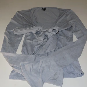 Grå tröja från Gina Tricot - Säljer en stilren knallblå tröja från Gina Tricot. Toppen har långa ärmar och ett knytband där fram och har en slits i mitten på magen. Perfekt för en chic och bekväm look.