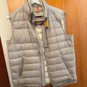 Ljusgrå dunväst från Parajumpers - Säljer en stilren ljusgrå dunväst från Parajumpers. Västen har en dragkedja framtill och en hög krage med knappdetalj. Västen är i nyskick med qr cod och kostar 3500 som ny.