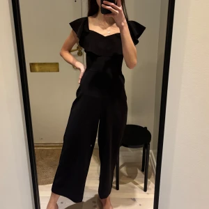 Svart jumpsuit med volangdetaljer - Snygg svart jumpsuit med volangdetaljer vid axlarna och en v-ringad design. Perfekt för en stilren look. 