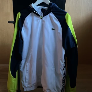 Vit och mörkblå vindjacka från Lacoste - Säljer en snygg vit och mörkblå vindjacka från Lacoste med neongula detaljer. Jackan har en dragkedja framtill och en huva. Perfekt för sportiga aktiviteter eller en casual look. Lacoste-loggan syns på bröstet och längs sidan.