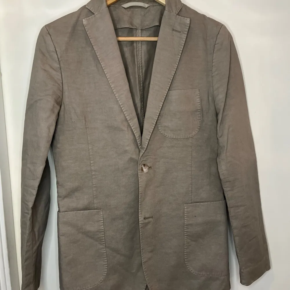 Snygg beige kavaj från J.Lindeberg i bomull. Perfekt för en stilren look med klassiska slag och fickor fram. Passar utmärkt till både formella och avslappnade tillfällen.. Puvut & Bleiserit.