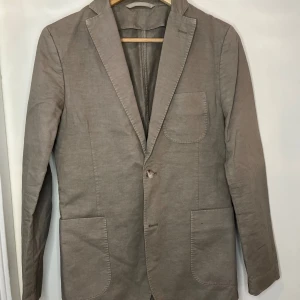 Beige kavaj från J.Lindeberg  - Snygg beige kavaj från J.Lindeberg i bomull. Perfekt för en stilren look med klassiska slag och fickor fram. Passar utmärkt till både formella och avslappnade tillfällen.