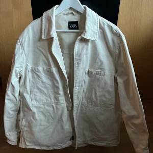 Vit overshirt från Zara - Säljer en stilren vit overshirt från Zara. Jackan har en klassisk krage och knappar framtill. Den är tillverkad i ett mjukt bomullsmaterial och har två stora fickor på framsidan. Perfekt för lager-på-lager-stilen.