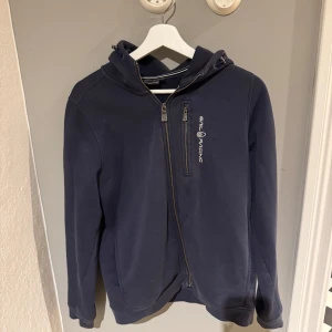 Mörkblå huvtröja från Sail Racing - Säljer en mörkblå huvtröja från Sail Racing med dragkedja framtill och logga på bröstet. Tröjan har en bekväm huva och långa ärmar, perfekt för kyligare dagar. Stilren och enkel design som passar till det mesta.