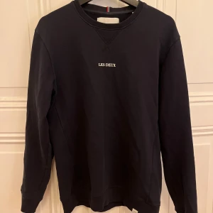 Mörkblå sweatshirt från Les Deux - Mörkblå sweatshirt från Les Deux med logga mitt på bröstet. Tröjan har långa ärmar och en ribbad detalj vid halsringningen. 
