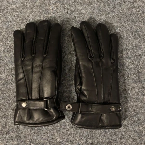 Svarta läderhandskar med foder - Säljer dessa eleganta svarta läderhandskar med mjukt foder och justerbar rem med knappdetalj vid handleden. Perfekta för att hålla händerna varma under kalla vinterdagar. Stilren design som passar till det mesta. 🖤UF-Företag så vi har från storlek S-L
