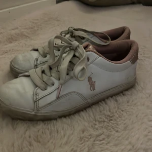 Vita sneakers med glittrig detalj från Ralph Lauren -  Snygga vita sneakers från Ralph Lauren med glittriga detaljer i rosa på hälen. Skorna har snörning och en klassisk logga på sidan. Perfekta för en stilren look. 💞