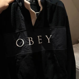 Svart fleece från Obey - Säljer en svart fleece hoodie från Obey med dragkedja och stort tryck på bröstet. Tröjan har en mysig insida och är perfekt för kyliga dagar. Den har en klassisk passform och är tillverkad i mjukt material. Storlek xs