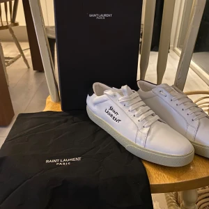 Vita sneakers från Saint Laurent   - Snygga vita sneakers från Saint Laurent med svart detalj på hälen. Skorna har klassisk snörning och en stilren design med Saint Laurent-logga på sidan. Perfekta för en trendig och avslappnad look.