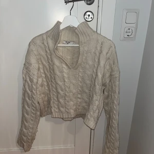 Beige stickad tröja från NA-KD - Säljer en snygg beige stickad tröja från NA-KD. Tröjan har en hög krage och ett kabelstickat mönster. Längst upp i kragen finns ett spänne om man vill ta ihop den i halsen💕