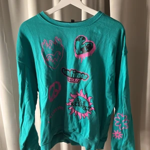 Grön tröja med tryck - Cool grön tröja med olika tryck i rosa och svart, inklusive hjärtan och text. Tröjan har långa ärmar och en avslappnad passform. Perfekt för en avslappnad stil med en färgglad touch.