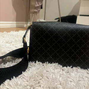 Svart axelväska med mönster - Snygg svart axelväska från By Malene Birger med ett diskret rutigt mönster. Väskan har en justerbar axelrem och en praktisk insida med flera fack. Perfekt för att bära med sig det viktigaste med stil.