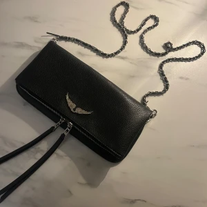 Svart axelväska med kedja och vingdetalj - Snygg svart axelväska i skinn med en cool kedjerem och en unik vingdetalj i silver på framsidan. Väskan har en dragkedja för säker förvaring och extra fack på baksidan. Perfekt för att ge din outfit en edgy touch.