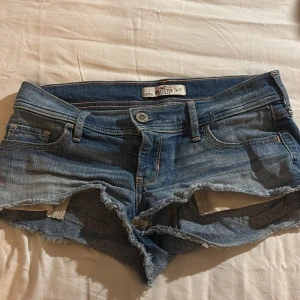Blå jeansshorts från Hollister - Säljer ett par blå jeansshorts från Hollister med fransiga kanter och klassisk femficksdesign. Perfekta för en avslappnad stil. De har en knapp och dragkedja framtill.