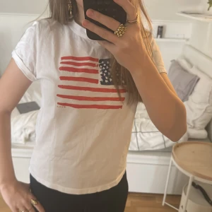 Vit t-shirt med amerikansk flagga - Säljer en vit t-shirt med tryck av den amerikanska flaggan i rött och blått. T-shirten har en klassisk rund hals och korta ärmar. Perfekt för en avslappnad stil. Hör av dig vid intresse! 🇺🇸