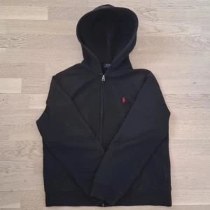 Stilren Ralph Lauren Hoodie - Svart, zip, XS - Letar du efter en klassisk och tidlös hoodie? Denna svarta Ralph lauren zip-hoodie i storlek XS är perfekt för både avslappnande och mer stiliga outfits. Den är i bra skick och har en bekväm passform. Med den ikoniska Ralph Lauren-loggan på bröstet och kvalitetsmaterial, är den både slitstark och bekväm. Perfekt för lager-på-lager-styling eller som en lätt jacka på våren. Passar dig som gillar en snygg, casual look med ett premium märke!