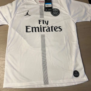 Vit PSG fotbollströja med Neymar JR - Säljer en vit PSG fotbollströja med Neymar JR och nummer 10 på ryggen. Tröjan har korta ärmar och Fly Emirates-logga på framsidan. Jordan-logga och PSG-emblem pryder bröstet. Perfekt för fotbollsfans!