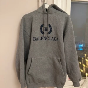 Grå hoodie från Balenciaga - Säljer en stilren grå hoodie från Balenciaga med blå logotyp på framsidan. Tröjan har en klassisk design med känguruficka och justerbar huva. Perfekt för en avslappnad look.