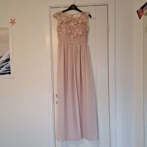 Ljusrosa långklänning med blommig spets - Elegant ljusrosa långklänning med vacker blommig spetsdetalj på överdelen. Klänningen har en romantisk och flytande design, perfekt för speciella tillfällen. Den är ärmlös och har en mjuk och feminin look.