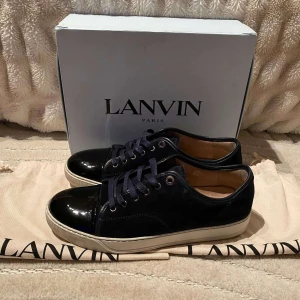 Svarta sneakers från Lanvin - Snygga svarta sneakers från Lanvin med glansig tå och snörning. Skorna har en vit sula och är perfekta för en stilren look. Levereras med originalförpackning och dustbag.