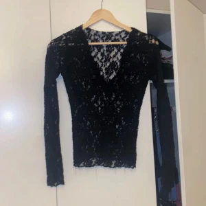 Svart spetsblus från Gina Tricot - Säljer en elegant svart spetsblus från Gina Tricot. Blusen har en v-ringad design och långa ärmar, perfekt för en stilfull look. Den är tillverkad i ett vackert spetsmaterial som ger en feminin touch.