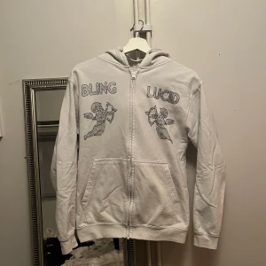 Vit hoodie från Blinglucid - Säljer en vit hoodie från Blinglucid med dragkedja. Den har ett tryck av änglar och texten 'BLING LUCID' på framsidan.  