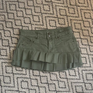 Grön kjol med volang, Gina  - Snygg grön kjol från Gina med volangdetalj, omsydd från jeans. Kjolen är låg i midjan och är tillverkad i ett jeansliknande material. Den har praktiska fickor och knappar framtill för en cool look.