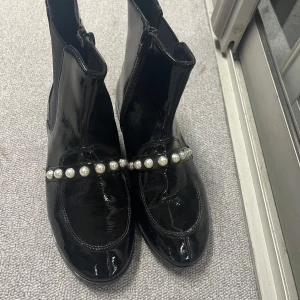 Svarta boots med pärldetaljer från Zara - Snygga svarta boots i lack från Zara med pärldetaljer över foten. De har en dragkedja på sidan för enkel på- och avtagning. Perfekta för att ge en elegant touch till din outfit.