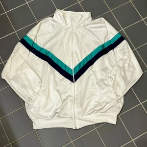 Vit vindjacka från Stinna - Säljer en vit vindjacka från Stinna med dragkedja och snygga diagonala ränder i turkos och mörkblått. Jackan har en hög krage och ribbade muddar vid ärmslut och nederkant. 