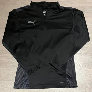 Svart långärmad tröja från Puma - Säljer en svart långärmad träningströja från Puma. Tröjan har en stilren design med ett snyggt mönster lvis sidorna. Gjord av syntetmaterial, perfekt för träning.