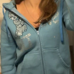 Von studio kofta - Säljer en blå hoodie med dragkedja och glittriga detaljer. Den har en tryckt figur på bröstet och långa ärmar. Perfekt för en avslappnad stil. 