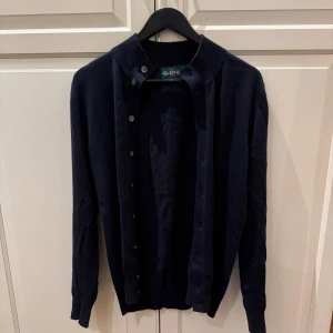 Mörkblå kofta från Alan Paine - Säljer en stilren mörkblå kofta från Alan Paine i meninoull. Koftan har långa ärmar och knappar framtill, perfekt för en elegant look. 