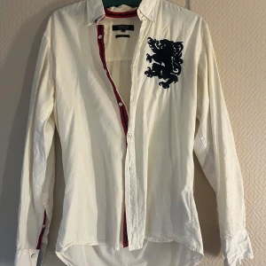 Vit skjorta med lejonbroderi från SMOG - Snygg vit skjorta från SMOG med ett coolt svart lejonbroderi på bröstet. Skjortan har en slim passform och långa ärmar. Den har röda och svarta detaljer längs knappslån och kragen, vilket ger en extra touch av stil.