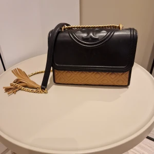 Svart och beige axelväska med kedja Tory burch  - Snygg axelväska i svart och beige med quiltad design. Väskan har en elegant guldkedja och en dekorativ tofs. Perfekt för att ge din outfit en stilfull touch.