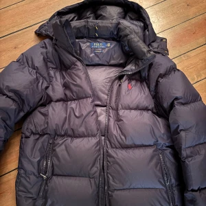 Mörkblå dunjacka från Polo Ralph Lauren - Säljer en snygg mörkblå dunjacka från Polo Ralph Lauren. Jackan har en dragkedja framtill och två praktiska fickor med dragkedjor. Den är perfekt för kyliga dagar och har en klassisk design med en liten röd logotyp på bröstet.