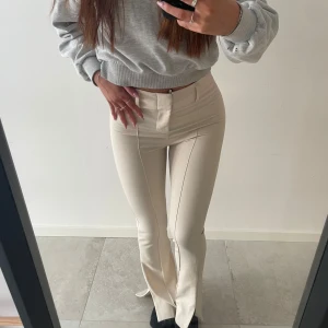 Beigea kostymbyxor  med slits - Snygga beige byxor med hög midja och slits vid anklarna. De har en tight passform och är perfekta för en stilren look. Byxorna har en dragkedja och knapp framtill.