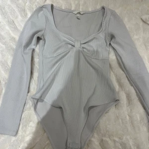 Ljusgrå ribbad bodysuit - Snygg ljusgrå bodysuit med ribbad struktur och långärmad design. Den har en elegant knytdetalj framtill och en djup, rundad halsringning. Perfekt för att skapa en stilren look. Dock har den en liten fläck under armen, men den syns inte så pass mycket att det stör