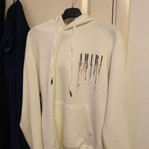 Vit hoodie från Amiri - Säljer en snygg vit hoodie från Amiri med färgglada droppdetaljer och logotyp på både fram- och baksidan. Den har en klassisk känguruficka och justerbar huva med snören. Perfekt för en stilren och modern look.