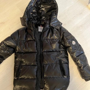  Moncler Maya jacka  - Snygg svart dunjacka från Moncler med glansig finish och huva. Jackan har dragkedja och praktiska fickor med knappar. Perfekt för kyliga dagar med stil.
