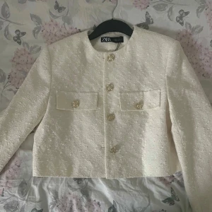 Vit kavaj från Zara - Säljer en elegant vit kavaj från Zara med en stilren design. Kavajen har långa ärmar och knappar framtill samt två fickor med lock. Perfekt för att ge en sofistikerad touch till din outfit. Helt ny och oanvänd.