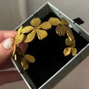Guldarmband med blommotiv - Elegant guldarmband med vackra blommotiv. Armbandet har en unik design med detaljerade blommor som ger en stilfull och sofistikerad look. Perfekt för att lägga till en touch av glamour till din outfit.